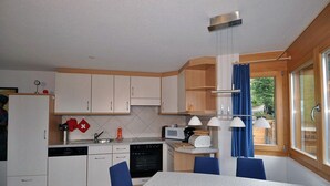 A compact kitchen with white cabinets, a dining table with blue chairs, and a window with blue curtains. - Camelot 4 (Bettmeralp)
