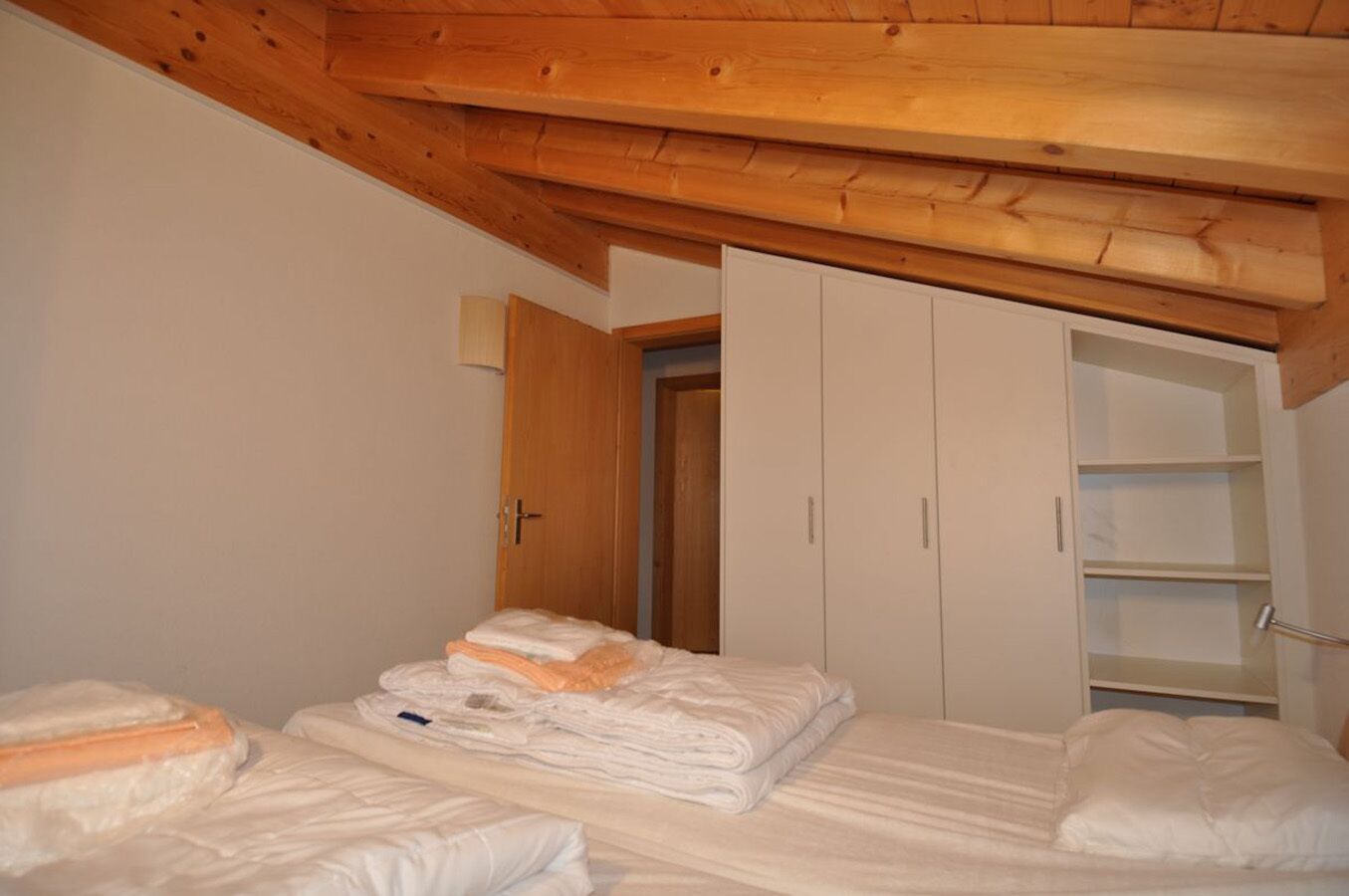 A bedroom with two beds, wooden ceiling beams, a wardrobe, and a door leading to another room.