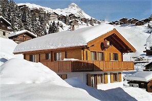 En træchalet med balkon, omgivet af sneklædte bjerge og andre chalet-lignende bygninger.