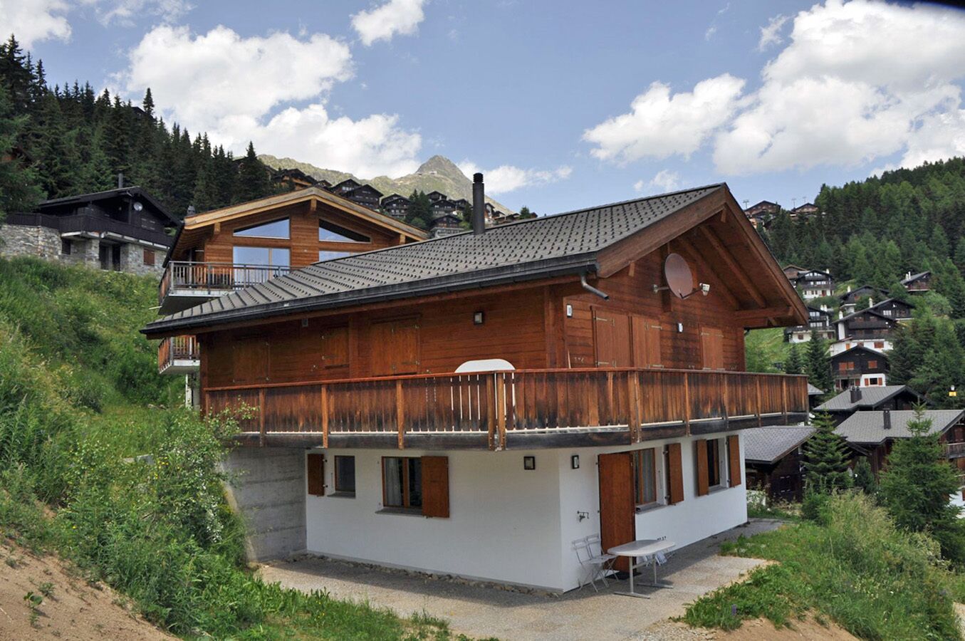 A wooden chalet with a balcony, white exterior walls, and a mountainous backdrop.
