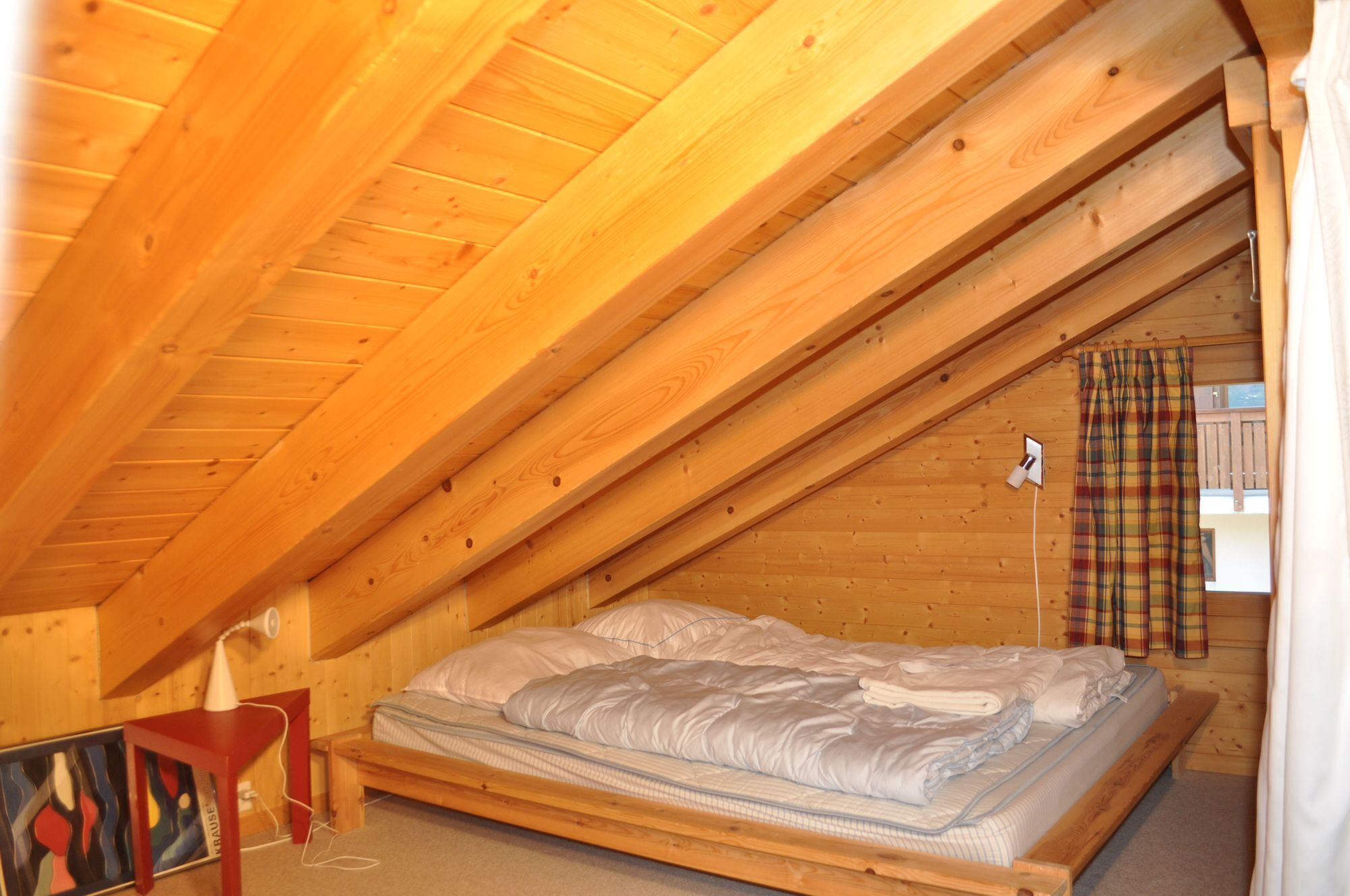 A small, cozy attic bedroom with a wooden sloped ceiling, a single bed, a small table, and a colorful painting on the wall.