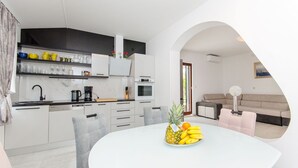 A modern kitchen with white cabinets, a dining table with a fruit bowl, and a living area with a sofa and a lamp. - Apartment Ana With Private Pool (Solin)