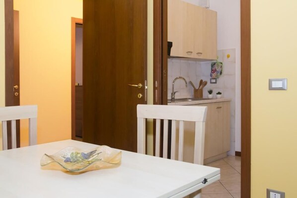 A room with a white table, a white chair, a wooden door, a kitchen area with cabinets and a sink.