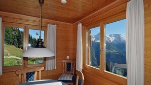 A wooden cabin room with a table, chairs, a hanging lamp, and large windows offering mountain views. - Harpa OG (Bettmeralp)