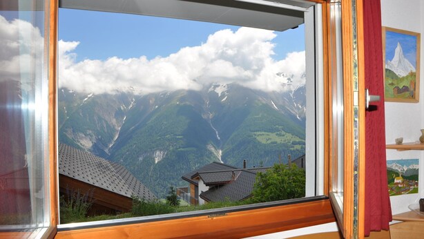 A window with a view of a mountainous landscape, including a house with a sloped roof and a mountain range with snow-capped peaks. - Heidi 16 (Bettmeralp)
