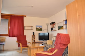 Un salón con un sofá rojo, una silla de madera, una mesita, un televisor y una estantería con libros y un cuadro enmarcado.