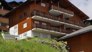 A multi-story wooden building with balconies, a satellite dish, and a small garden area with flowers and rocks. - Heidi 16 (Bettmeralp)