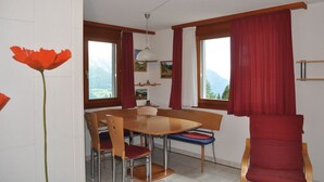 A dining area with a wooden table and chairs, a hanging light fixture, and large windows with red curtains. - Heidi 16 (Bettmeralp)