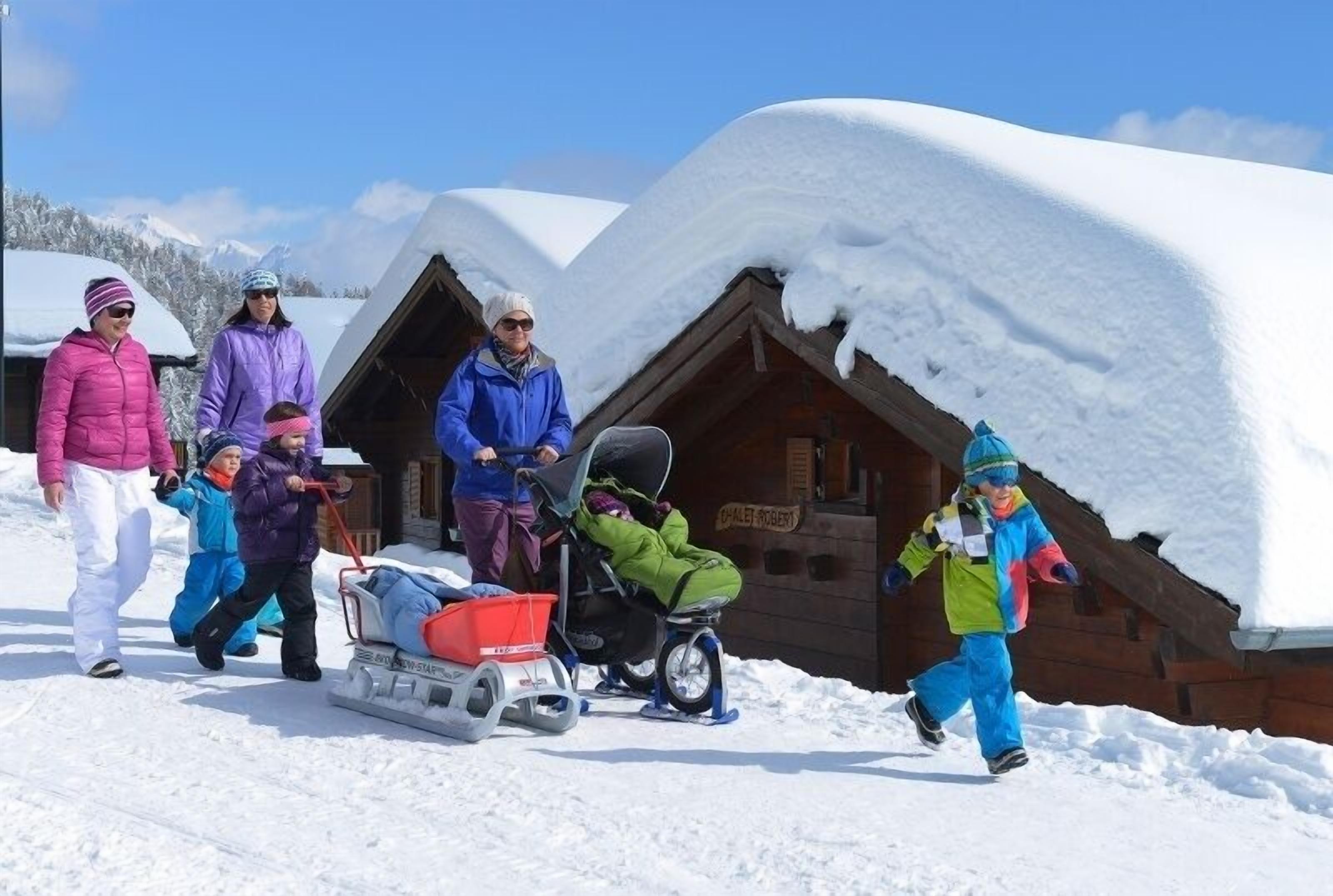 A group of people with a sled and a stroller in a snowy outdoor setting with wooden cabins in the background.