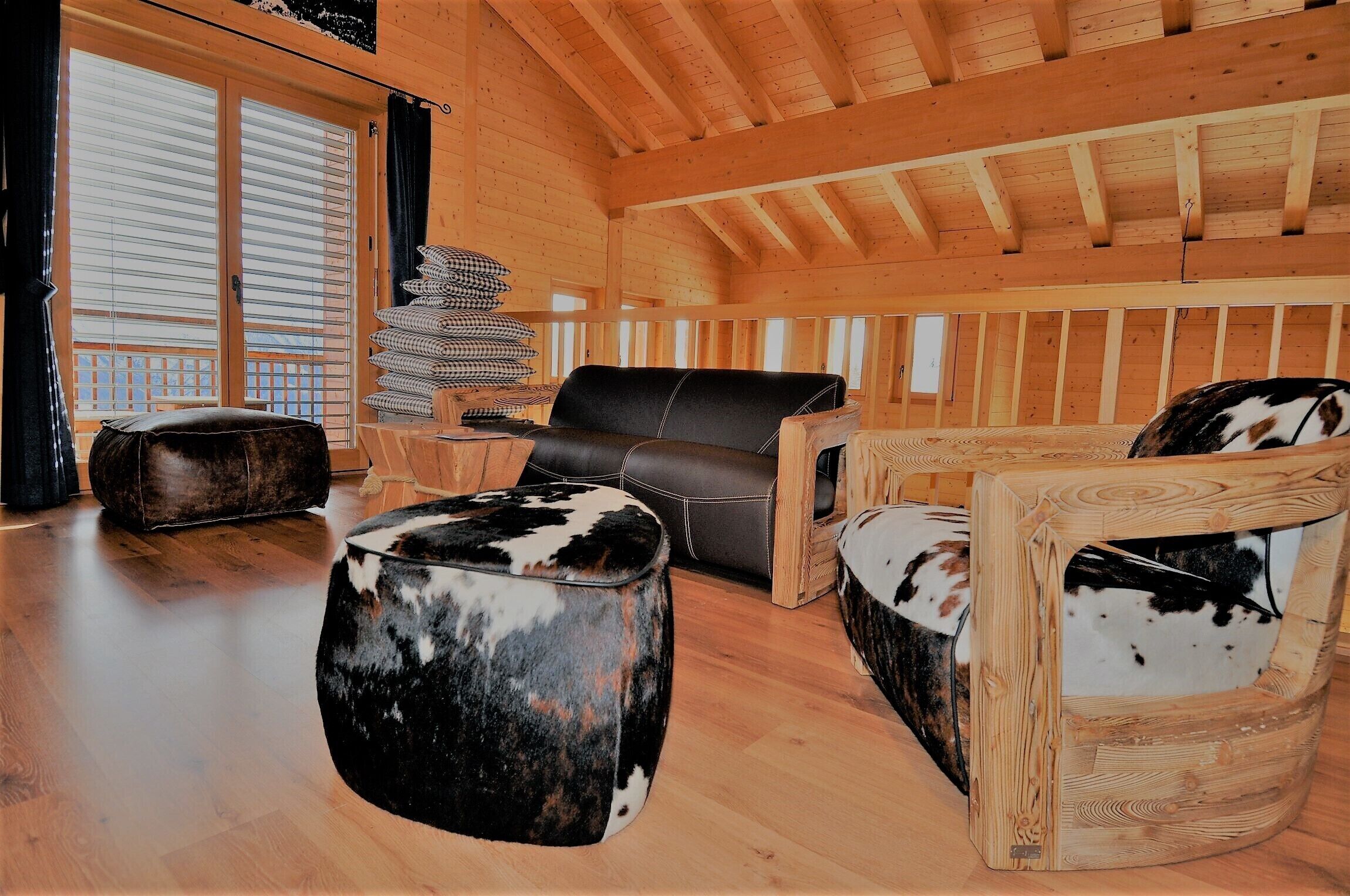 Ein gemütliches Holzhaus-Interieur mit einer Couch, Hocker und Sessel, einer Kissenstapel und einem Fenster mit Jalousien.