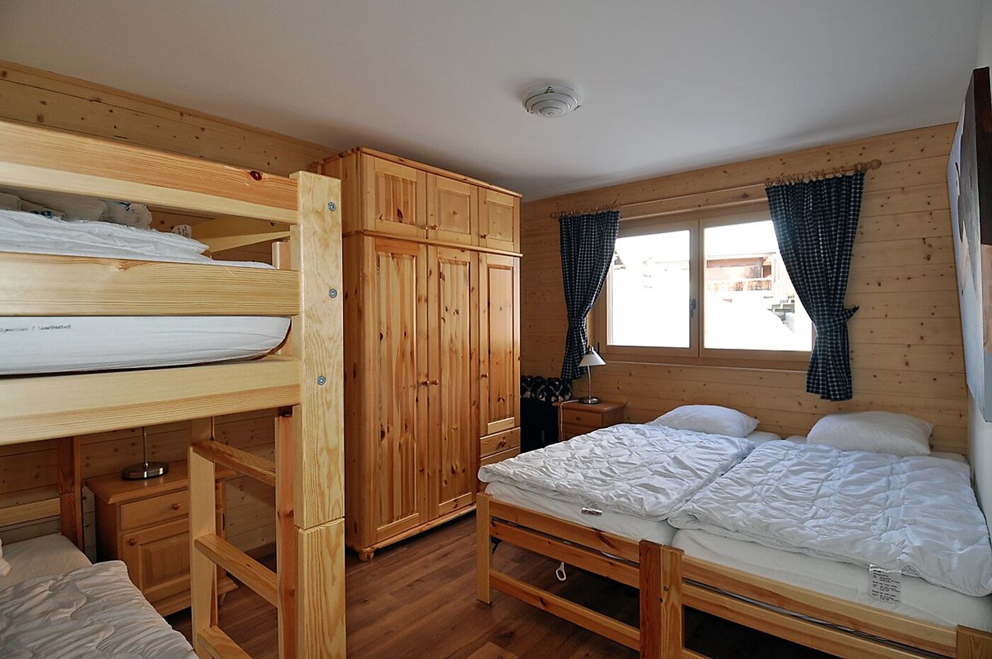 Een houten kamer met stapelbedden, een garderobe, een nachtkastje en een raam met gordijnen.