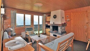 Un acogedor interior de cabaña con chimenea de piedra, muebles de madera y grandes ventanales con vistas a la montaña.