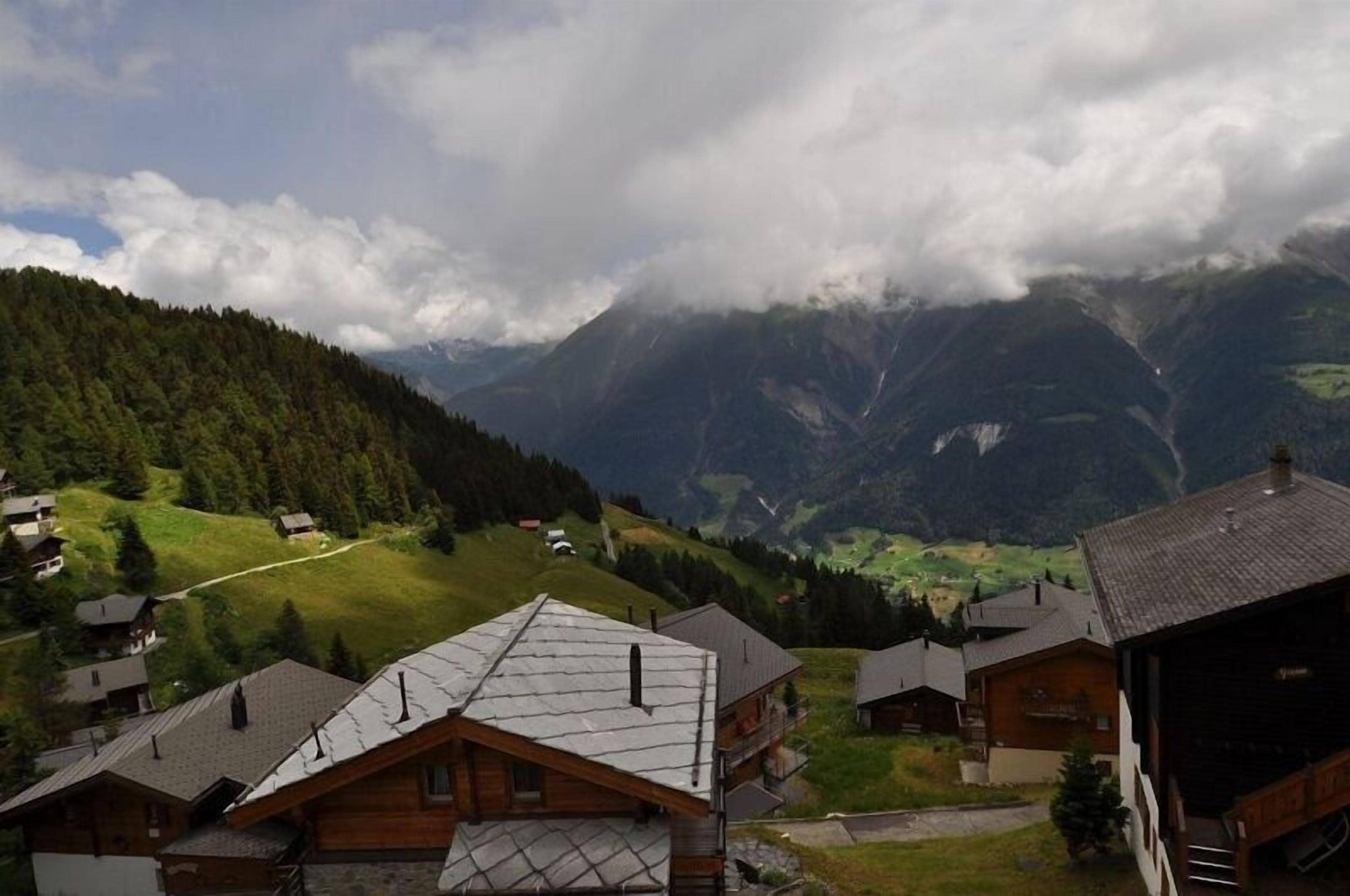 A mountain village with wooden houses, a winding road, and a valley with mountains in the background.