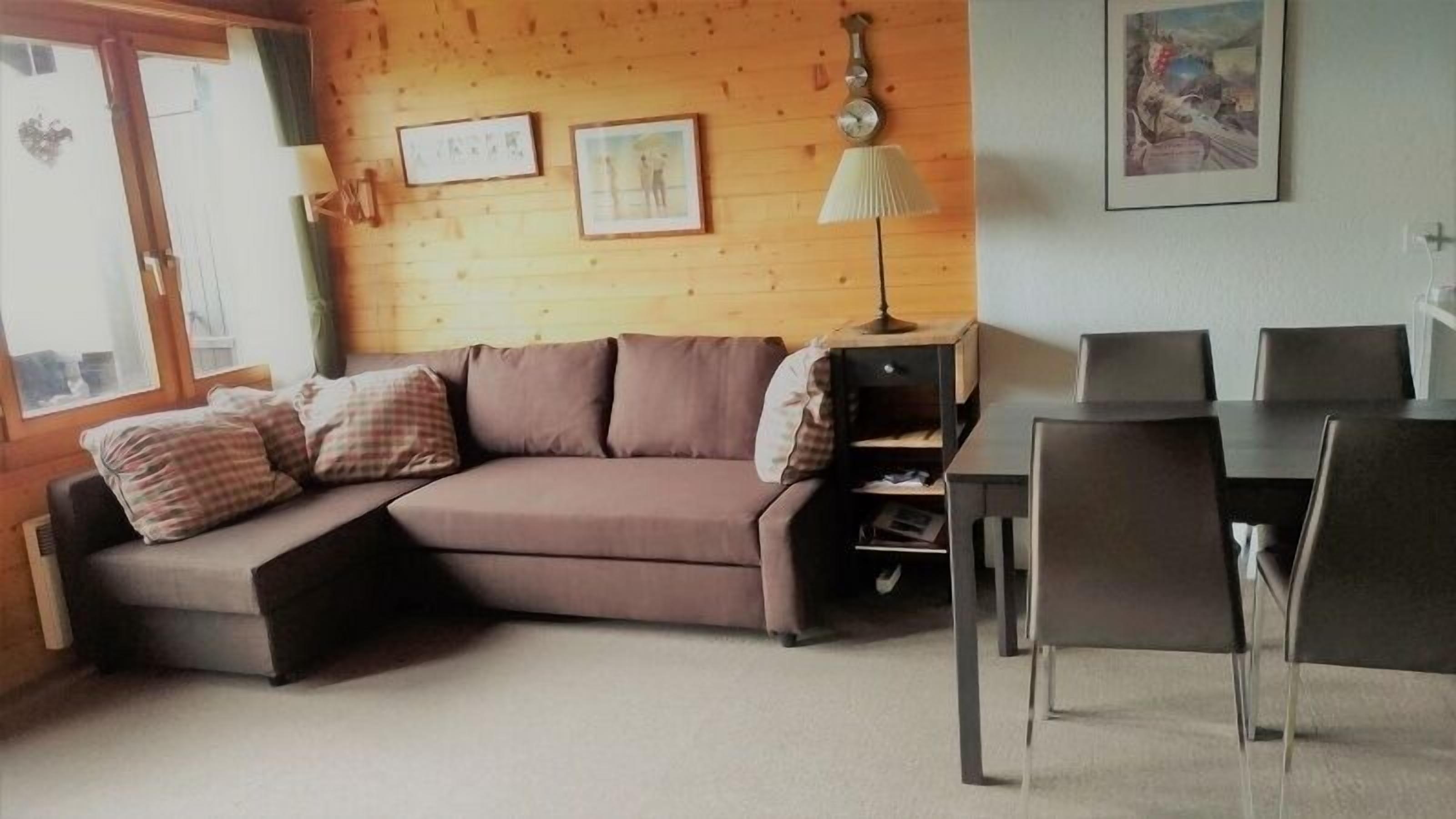 A cozy living room with a brown sofa, a dining table with chairs, and a lamp.