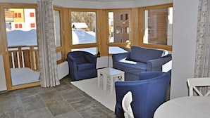 A room with a sofa, two armchairs, a small table, and large windows with a view of buildings and snow outside. - Mon Reve Bergh (Bettmeralp)