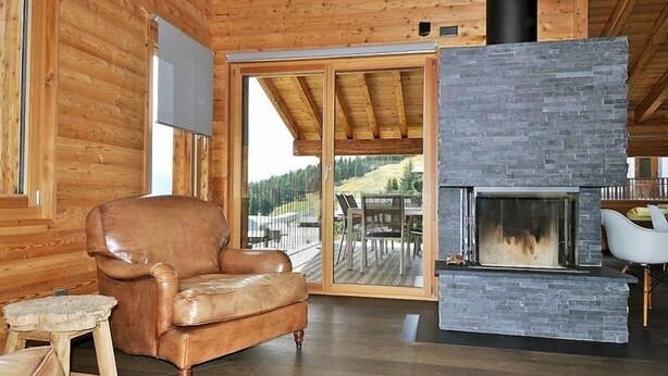 A cozy log cabin interior with a stone fireplace, leather armchair, and a view of a deck and forest. - Nachtigall OG (Bettmeralp)