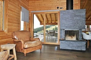 A cozy log cabin interior with a stone fireplace, leather armchair, and a view of a deck and forest.