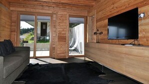 A modern living room with wooden walls, a flat-screen TV mounted on the wall, a sofa, and a large black rug. - Nachtigall OG (Bettmeralp)