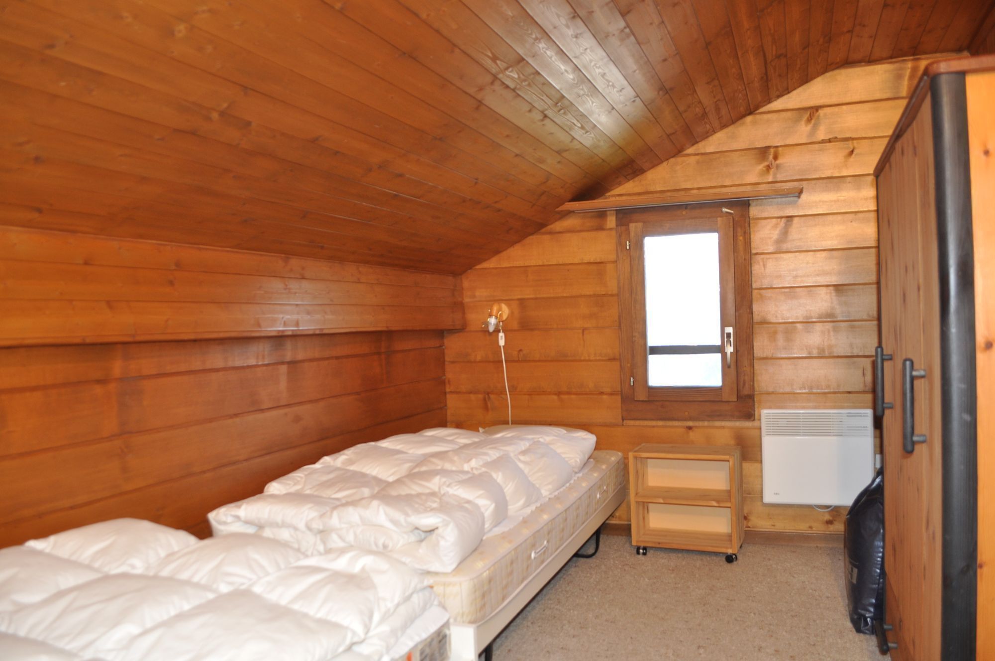 A small, wooden-paneled room with a single bed, a small table, and a window.