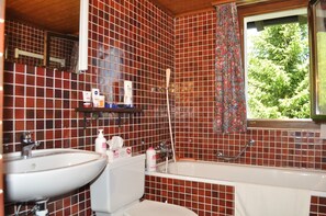 A bathroom with a white sink, toilet, and bathtub. There is a shelf with toiletries and a window with a view of greenery. - Polaris (Bettmeralp)