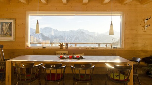 A wooden dining area with a long table and six chairs, a large window with a mountain view, and pendant lights. - Rupicapra (Bettmeralp)