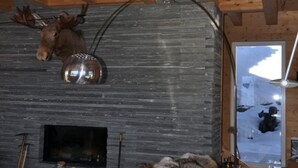 A rustic cabin interior with a stone fireplace, wooden beams, a mounted moose head, and a large window with a view of snow-covered mountains. - Rupicapra (Bettmeralp)