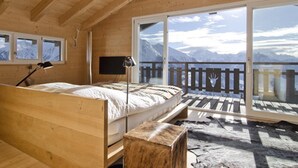 A wooden bedroom with a bed, a desk, and a large window overlooking mountains. - Rupicapra (Bettmeralp)
