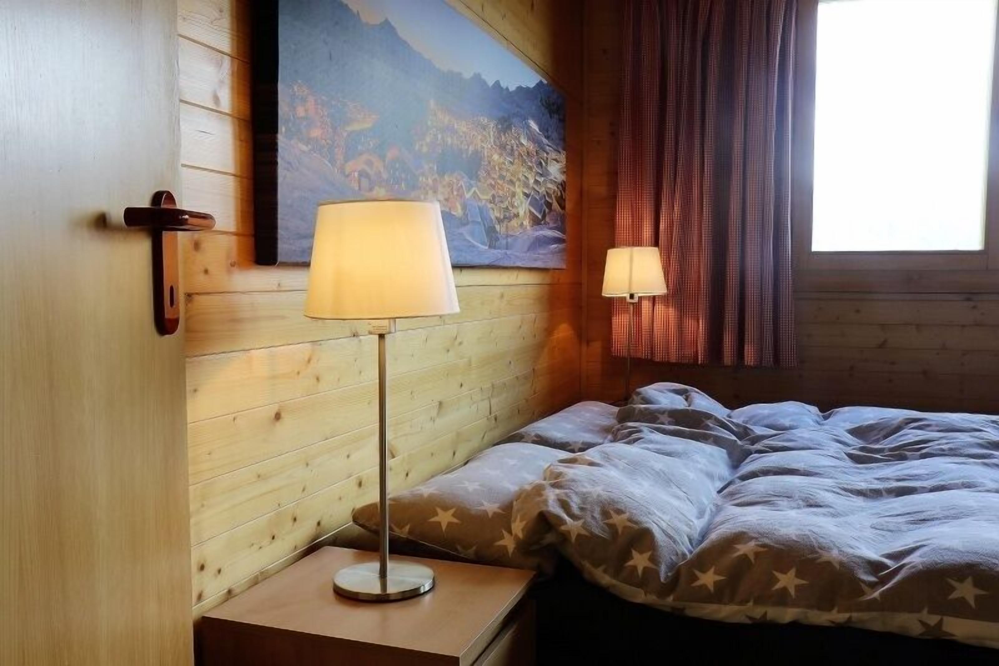 A wooden cabin room with a bed, a nightstand, a lamp, and a painting of a mountain landscape.