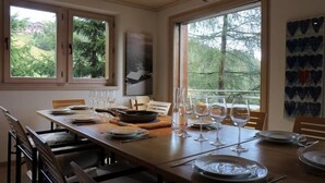 A dining area with a wooden table set for six, large windows with a view of trees, and a wall with abstract art. - Sternschnuppe OG (Bettmeralp)