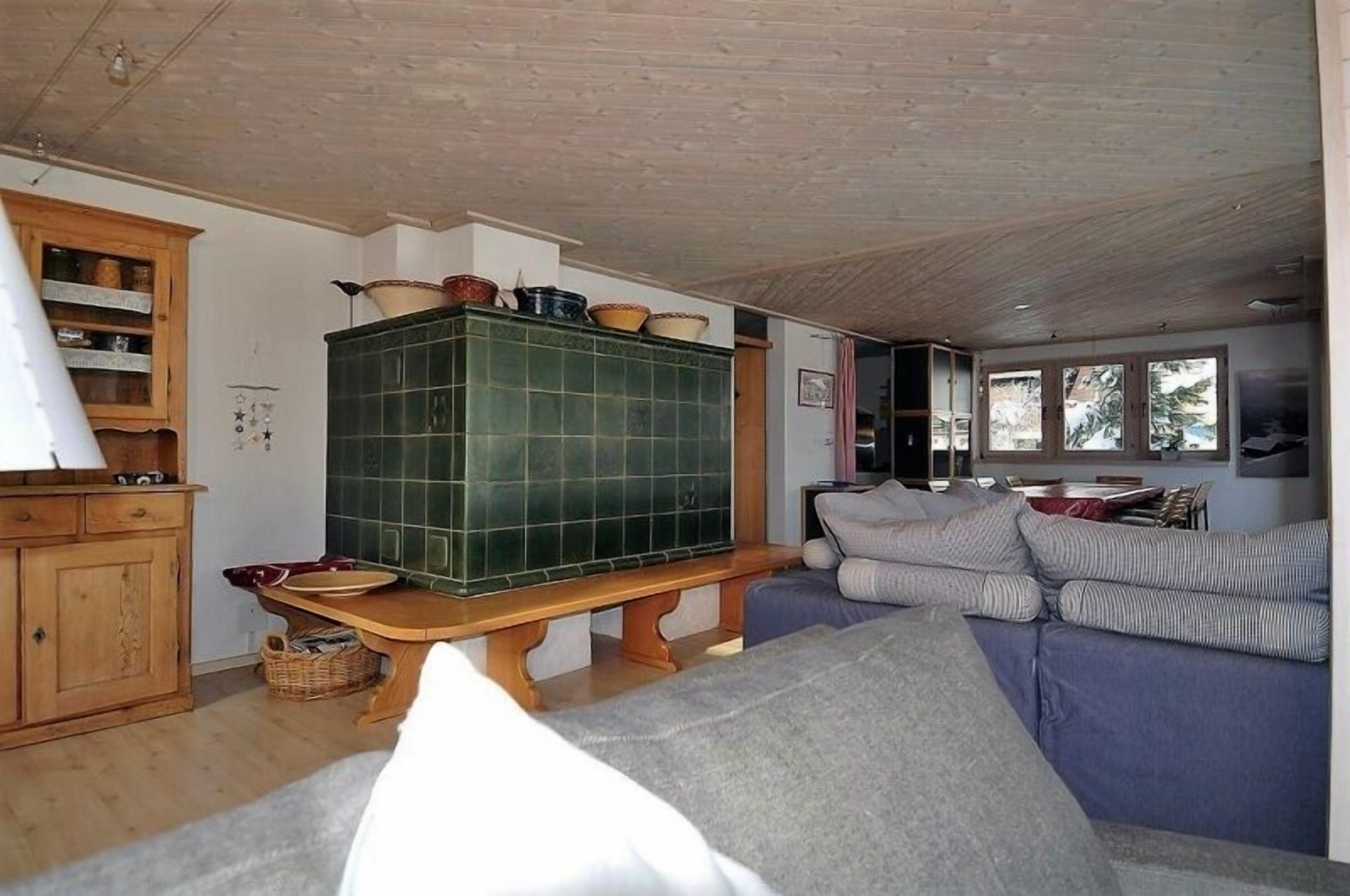 A cozy living room with a green tiled fireplace, wooden furniture, and a large window providing natural light.