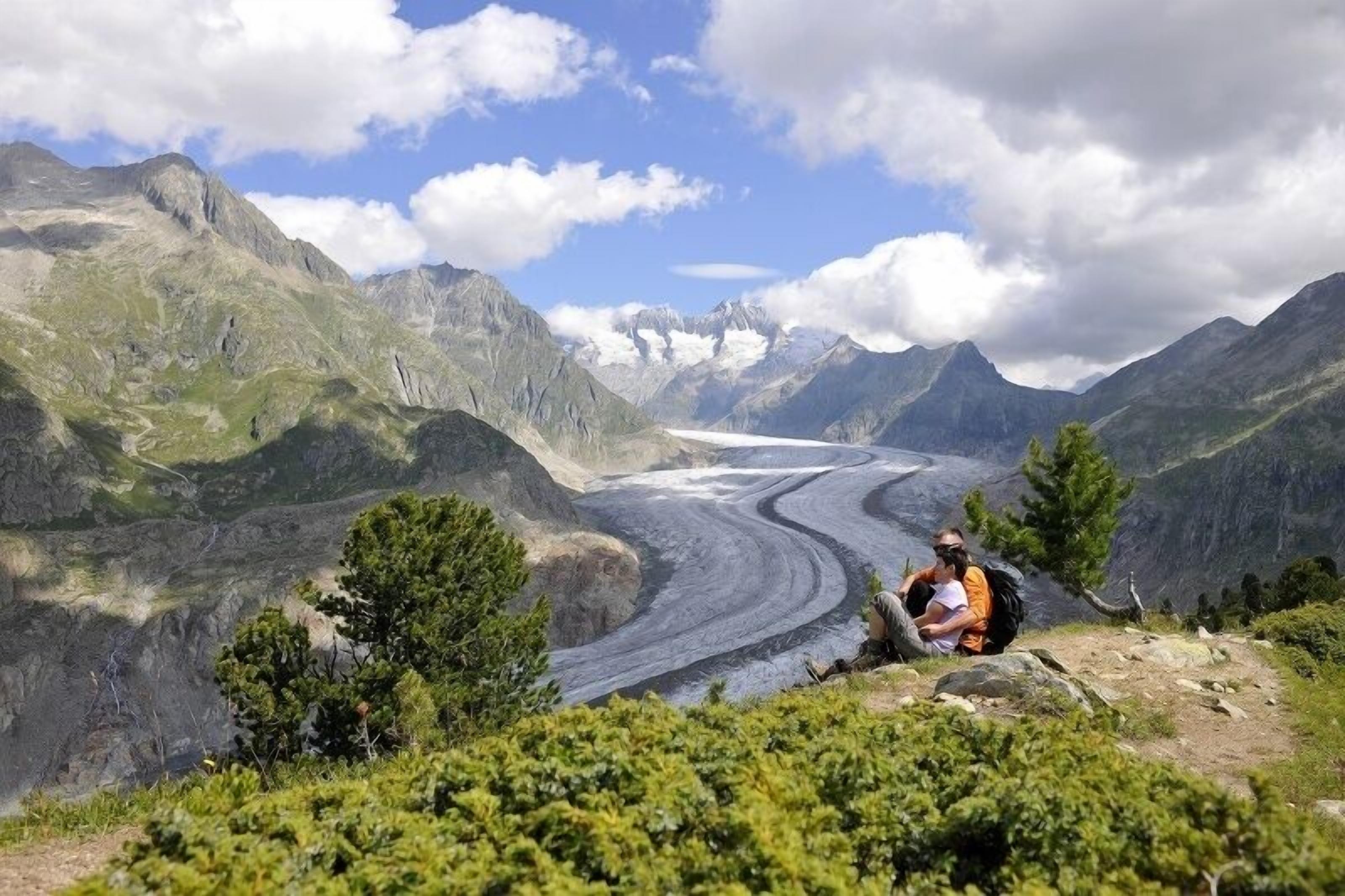 Paesaggio montuoso con un ghiacciaio, una persona seduta su una roccia e cielo sereno.
