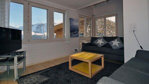 A modern living room with a flat-screen TV, a wooden coffee table, a sofa with star-patterned cushions, and large windows offering a mountain view. - Sternschnuppe OG (Bettmeralp)