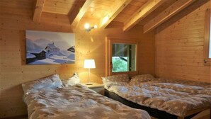 A cozy wooden cabin room with two beds, a painting of a snowy village, and a window with a view of greenery. - Sternschnuppe OG (Bettmeralp)