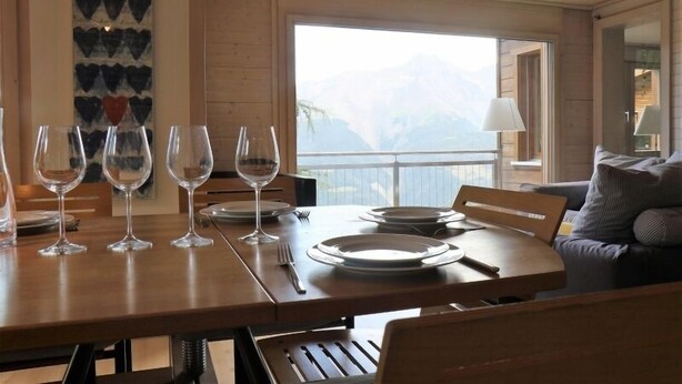 A dining area with a wooden table set for four, a view of mountains through a large window, and a modern interior design. - Sternschnuppe OG (Bettmeralp)