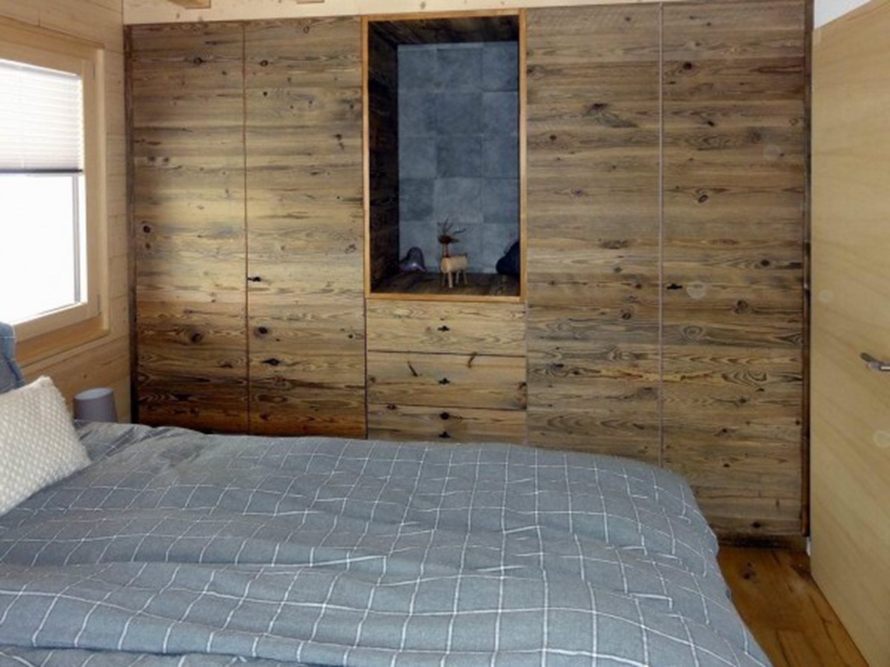 Un dormitorio con paneles de madera en la pared, una cama con colcha a cuadros grises y un ventanal pequeño con un cuadro enmarcado de un ciervo.