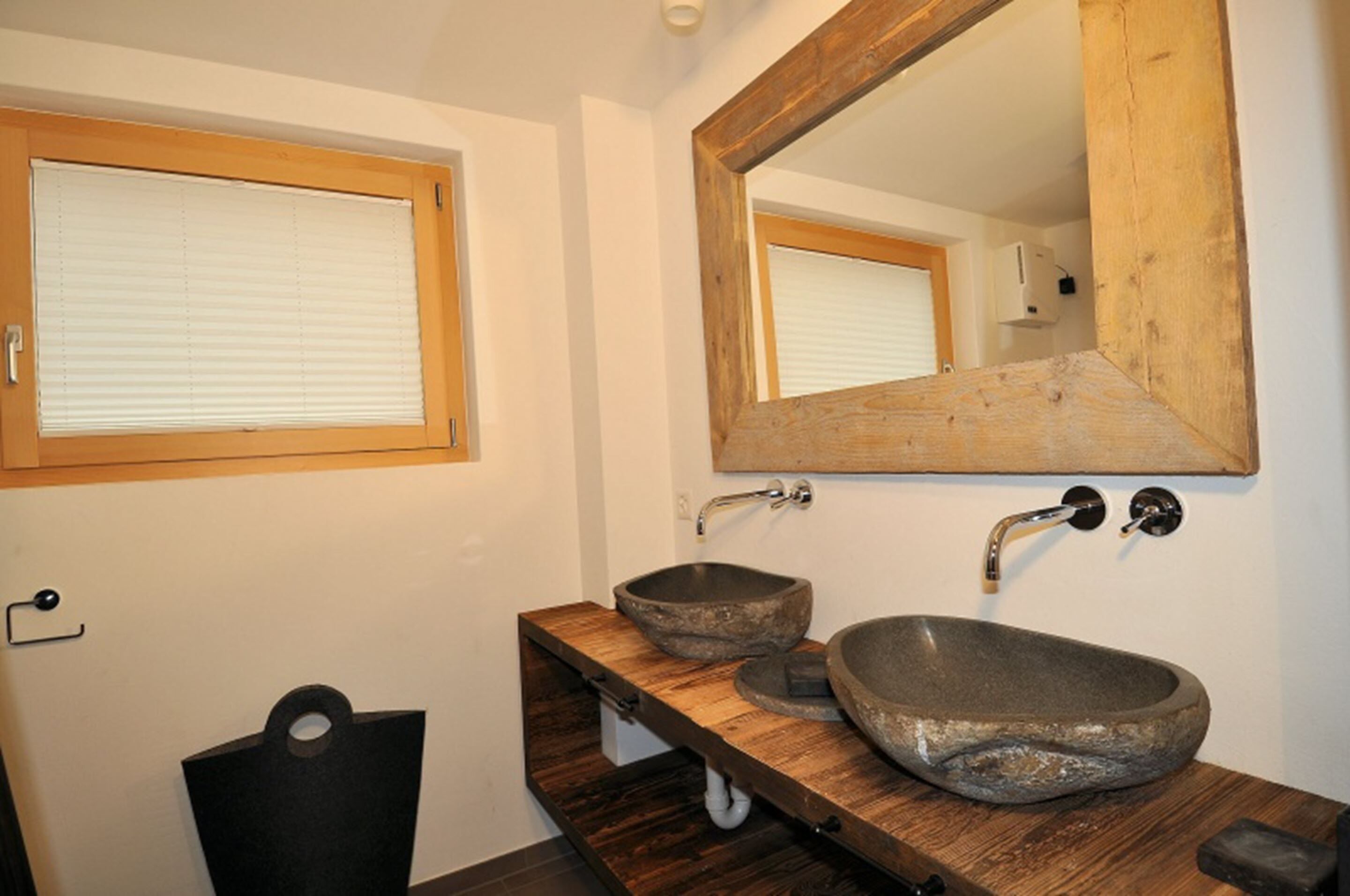 A bathroom with a wooden vanity, two stone sinks, a large mirror, and a window with blinds.