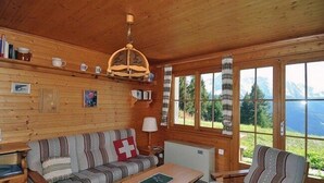 Interior acogedor de una cabaña con sofá, sillón, mesa, paredes de madera, ventana con vista a las montañas y una lámpara en el techo.