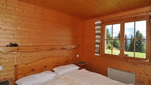 A wooden cabin room with two beds, a window with a view of mountains, and a shelf with a camera. - Toggi OG (Bettmeralp)