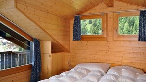 Une pièce de cabane en bois comprenant un lit, deux fenêtres avec des rideaux et une vue sur les arbres à l’extérieur.