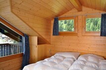 Habitación de cabaña de madera con una cama, dos ventanas con cortinas y vista a árboles.
