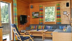 L’intérieur d’un chalet accueillant, avec un décor en bois, un canapé, un fauteuil à bascule, un téléviseur et une fenêtre agrémentée de rideaux.