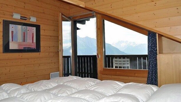 Une chambre avec un plafond en bois, un lit, une fenêtre donnant sur la montagne et un tableau encadré au mur.
