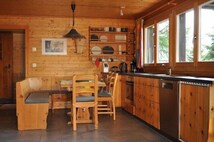 Interior de una cabaña de madera con comedor, cocina y ventanas amplias.