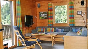 En mysig stuga med träpanel på väggarna, en soffa, en gungstol, en tv och ett fönster med gardiner.