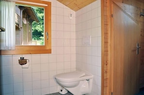 A small, clean bathroom with white tiles, a toilet, a window with a wooden frame, and a wooden door.