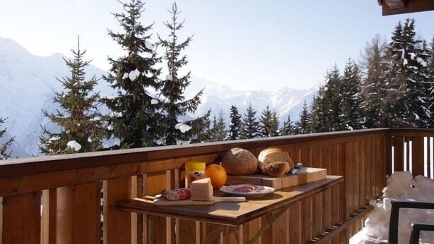 Een houten terras met een ontbijt tafel, met uitzicht op besneeuwde bergen en dennenbomen.