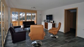 A spacious living area with a large window offering a mountain view, a black leather sofa, a brown leather armchair, and a television on a wooden cabinet. - Marjo OG (Ernen)