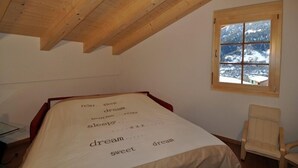 A bedroom with a wooden ceiling, a bed with a white sheet, a window with a view of snow-covered mountains, and a chair. - Marjo OG (Ernen)
