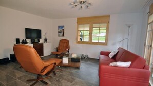 A living room with a red sofa, an orange chair, a glass coffee table, a TV, and a window with a view of greenery. - Marjo EG (Ernen)