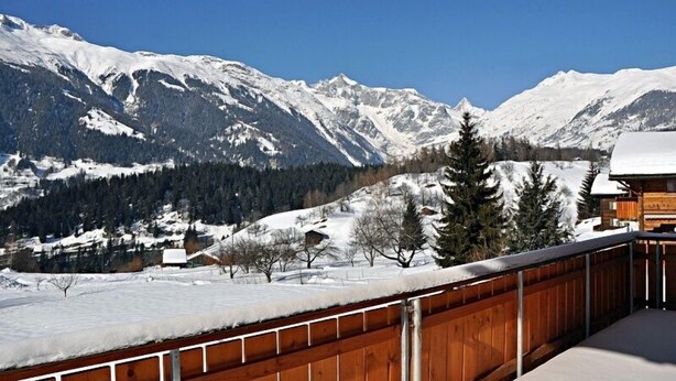 A wooden balcony with a view of snow-covered mountains and a forested valley. - Marjo EG (Ernen)